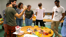 Un animateur présente à un groupe d'adultes différentes graines posées sur une table et une maracas fabriquée avec des graines et du carton