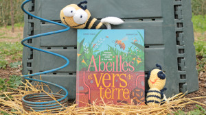 Un livre est posé devant un composteur avec deux abeilles en tricot posées dessus et à côté ainsi qu'un outil permettant d'aérer le compost