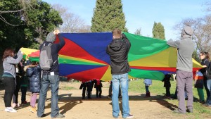 Des enfants inscrits à l'Accueil de loisirs de l'Écolothèque accompagnés de leurs parents jouent au jeu du parachute dans le grand pré
